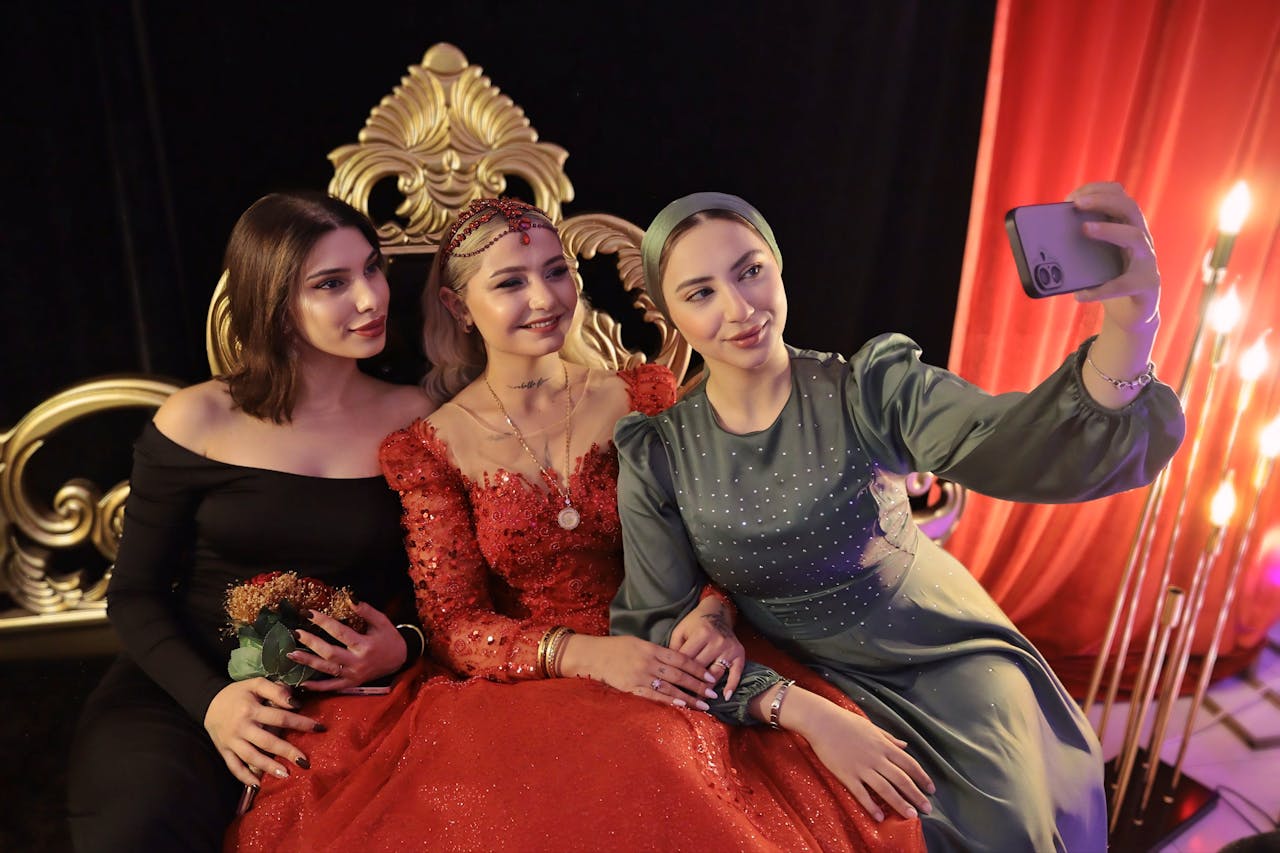 Three elegantly dressed women taking a selfie during a glamorous event with ornate decor.