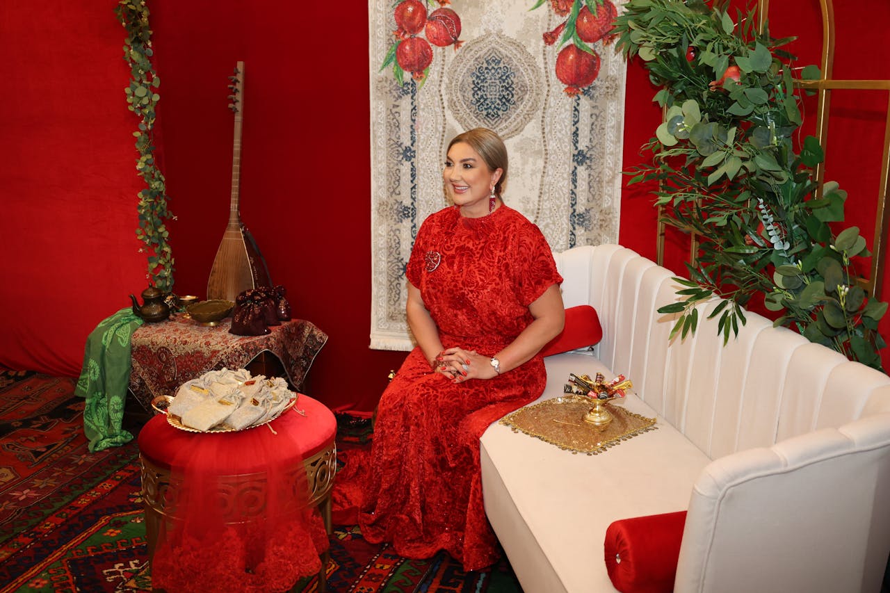 A woman in a vibrant red dress sits elegantly in a richly decorated room with cultural elements.