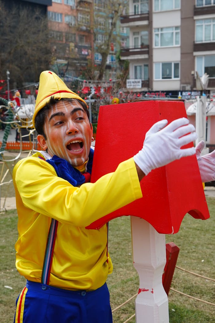 A cheerful clown in a vibrant costume embraces a red structure outdoors.