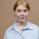 Close-up portrait of a young Caucasian woman wearing a blue shirt, neutral background.